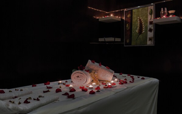 A dimly lit room with a bed covered in white sheets, surrounded by rose petals and lit candles. - Rest Night Hotel Suites Al Hamra (Riyadh)