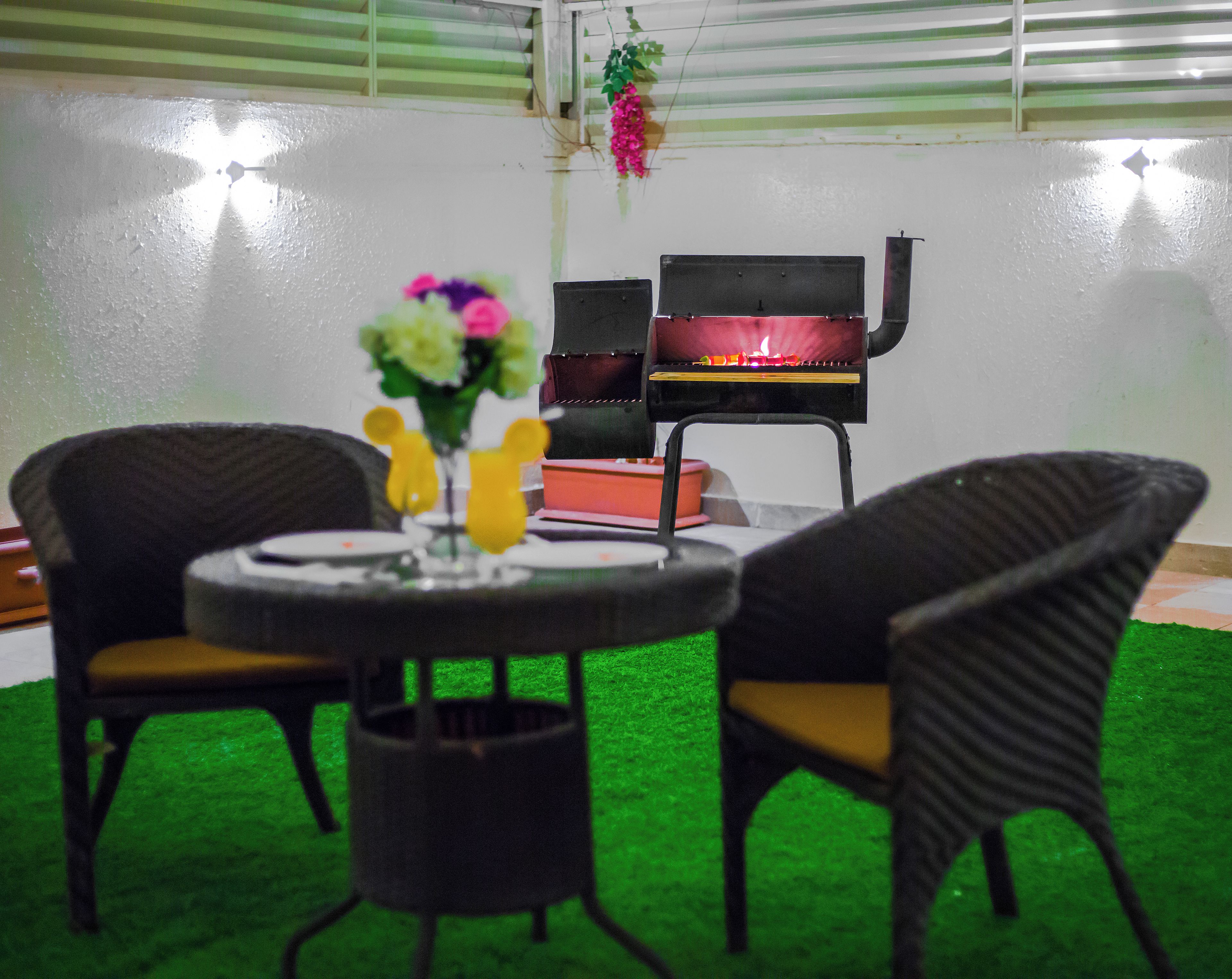 A room with a round table, two wicker chairs, a small vase with flowers, a grill, and a green carpet.