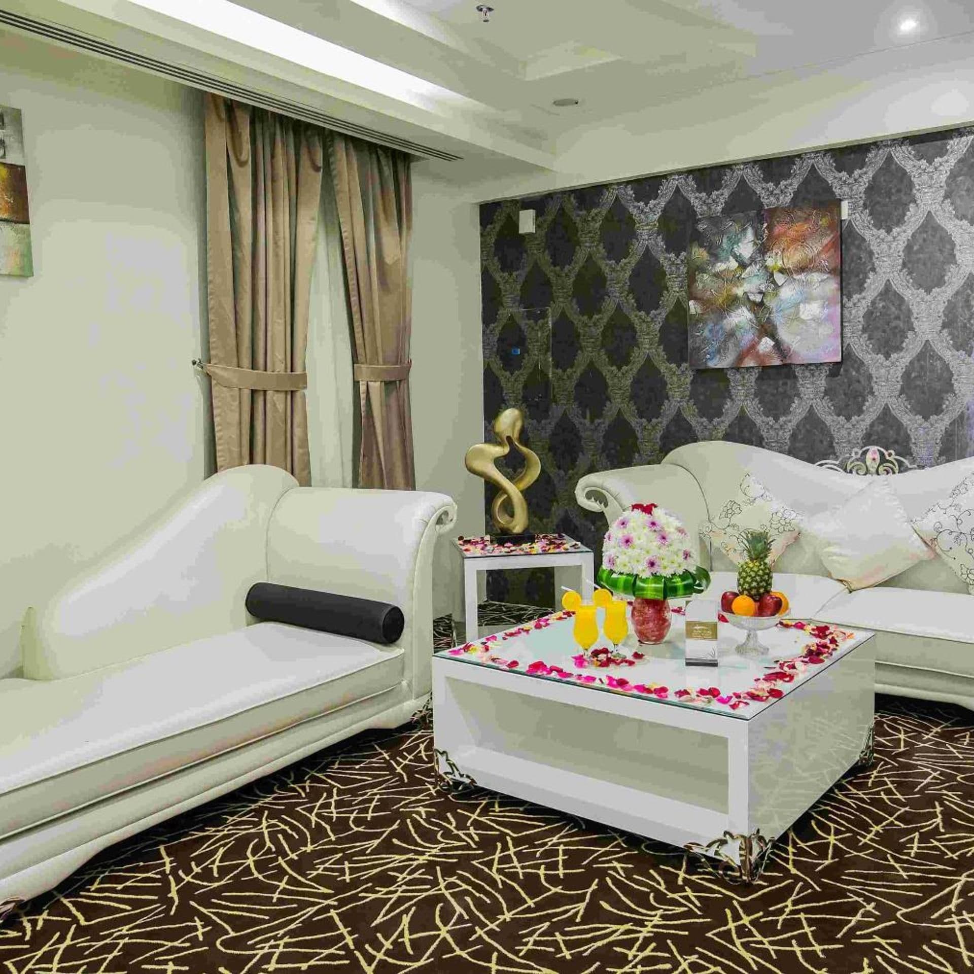 A living room with a white sofa, a coffee table with flowers and fruit, and a patterned wall.