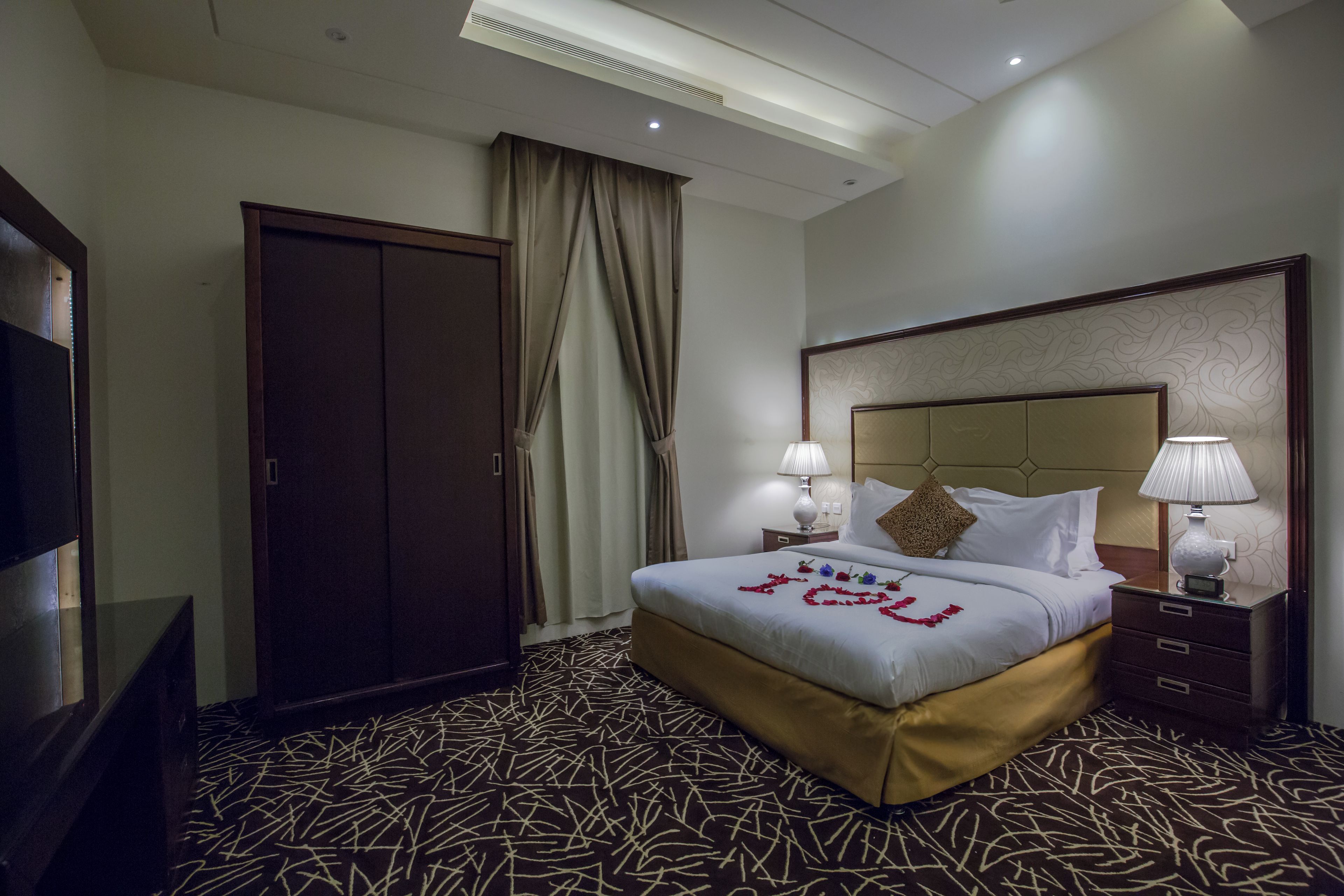 A hotel room with a large bed, a wardrobe, a television, and a nightstand with lamps.