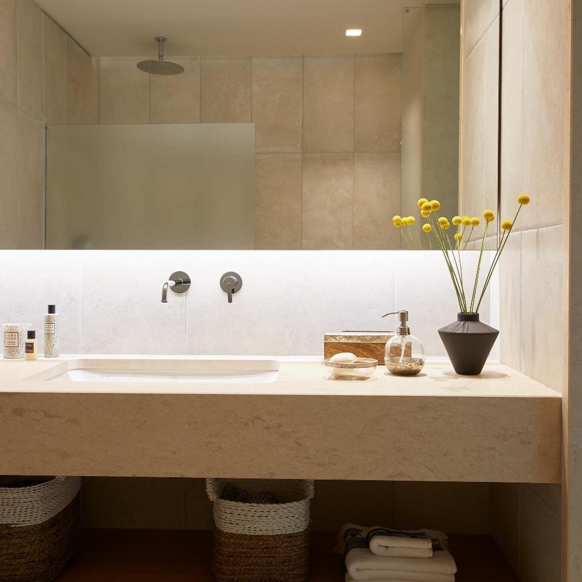 A bathroom with a sink, two faucets, a showerhead, a mirror, and a vase with yellow flowers.