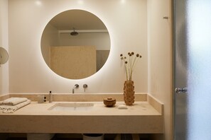 Ein Badezimmer mit rundem Spiegel, Waschbecken, Dusche und einer Vase mit getrockneten Blumen.