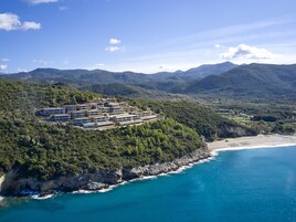 Un resort costero con varias edificaciones, una playa y terreno montañoso al fondo.