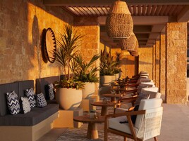 Un área de estar al aire libre iluminada por el sol, con paredes de piedra, plantas grandes en macetas y muebles de madera.