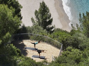A wooden deck with two individuals practicing yoga, overlooking a beach with clear water and surrounded by greenery. - Marbella Elix (Igoumenitsa)