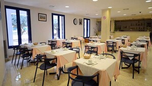 Un comedor con varias mesas y sillas, una cocina al fondo y ventanas amplias que dejan entrar luz natural.