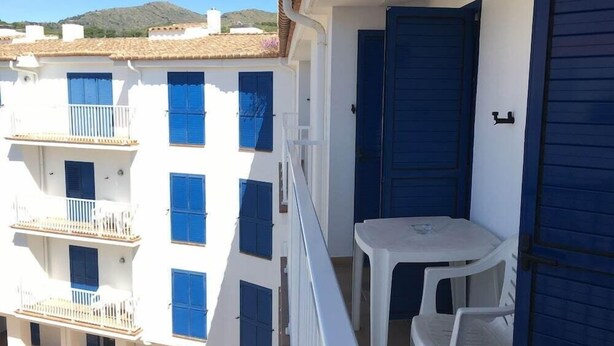 Un balcón con una mesa y sillas blancas, una puerta azul y un edificio blanco con contraventanas azules.