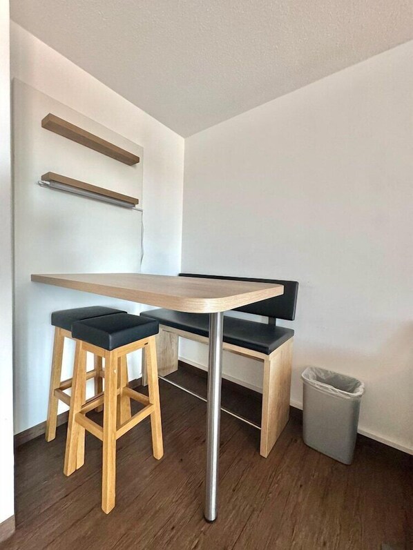 A modern, minimalist room with a wooden table and two black cushioned stools. There are two wooden shelves above the table and a trash bin to the right. - MY Parkhotel by AR Hotels (Kempten)