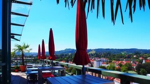 A terrace with tables and red umbrellas, overlooking a town with red-roofed buildings and greenery. - MY Parkhotel by AR Hotels (Kempten)