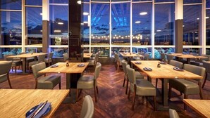 A restaurant with a modern interior, featuring wooden tables and chairs, large windows with a view of the cityscape, and a central pillar. - MY Parkhotel by AR Hotels (Kempten)
