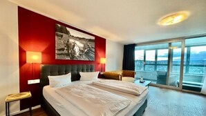 A hotel room with a large bed, a city view through floor-to-ceiling windows, and a red accent wall with a black and white artwork. - MY Parkhotel by AR Hotels (Kempten)