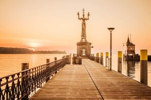 A wooden pier leading to a statue on a small island, with a lamp post and a tower on the right side. - Harbr Hotel Konstanz (Konstanz)