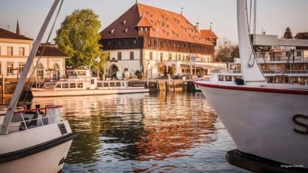 A marina with boats docked, a building with a red roof, and a tree in the background. - Harbr Hotel Konstanz (Konstanz)