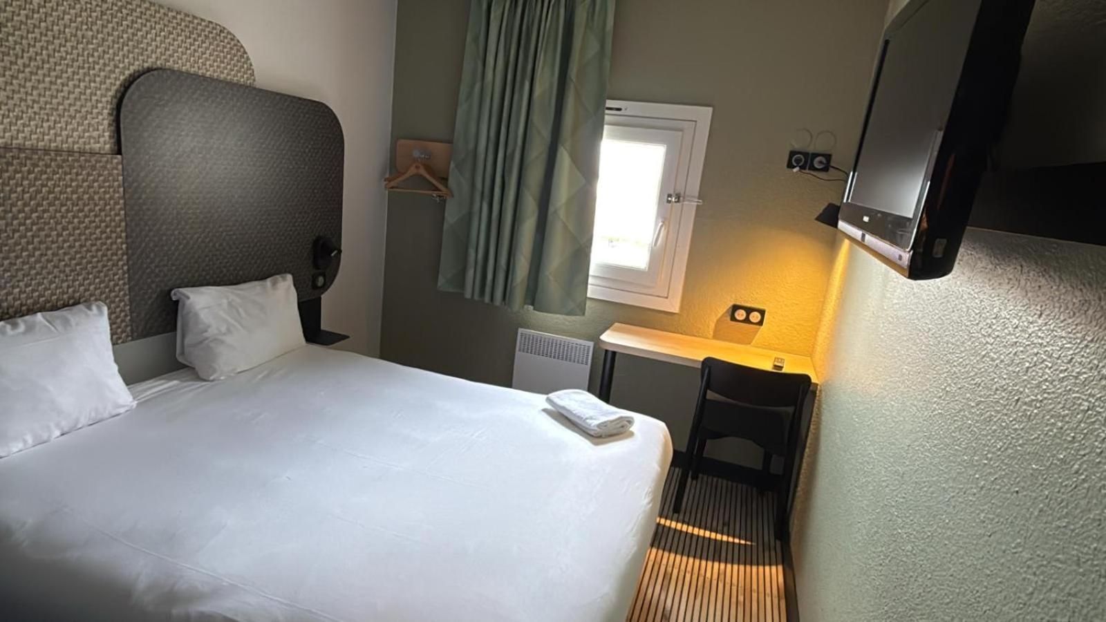 Habitación de hotel con una cama grande, un escritorio pequeño, una silla, una ventana con cortinas, un radiador y un televisor de pantalla plana montado en la pared.