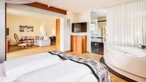 A modern hotel room with a large bed, a sitting area with a sofa and chairs, a TV, a glass-enclosed bathroom with a jacuzzi, and a wooden ceiling. - Romantik Hotel Im Weissen Roessl (Sankt Wolfgang im Salzkammergut)
