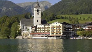 A lakeside building with a prominent tower, surrounded by lush greenery and mountains. - Romantik Hotel Im Weissen Roessl (Sankt Wolfgang im Salzkammergut)