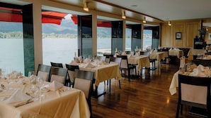A restaurant with tables set for dining, large windows offering a view of a lake and mountains, and wooden flooring. - Romantik Hotel Im Weissen Roessl (Sankt Wolfgang im Salzkammergut)