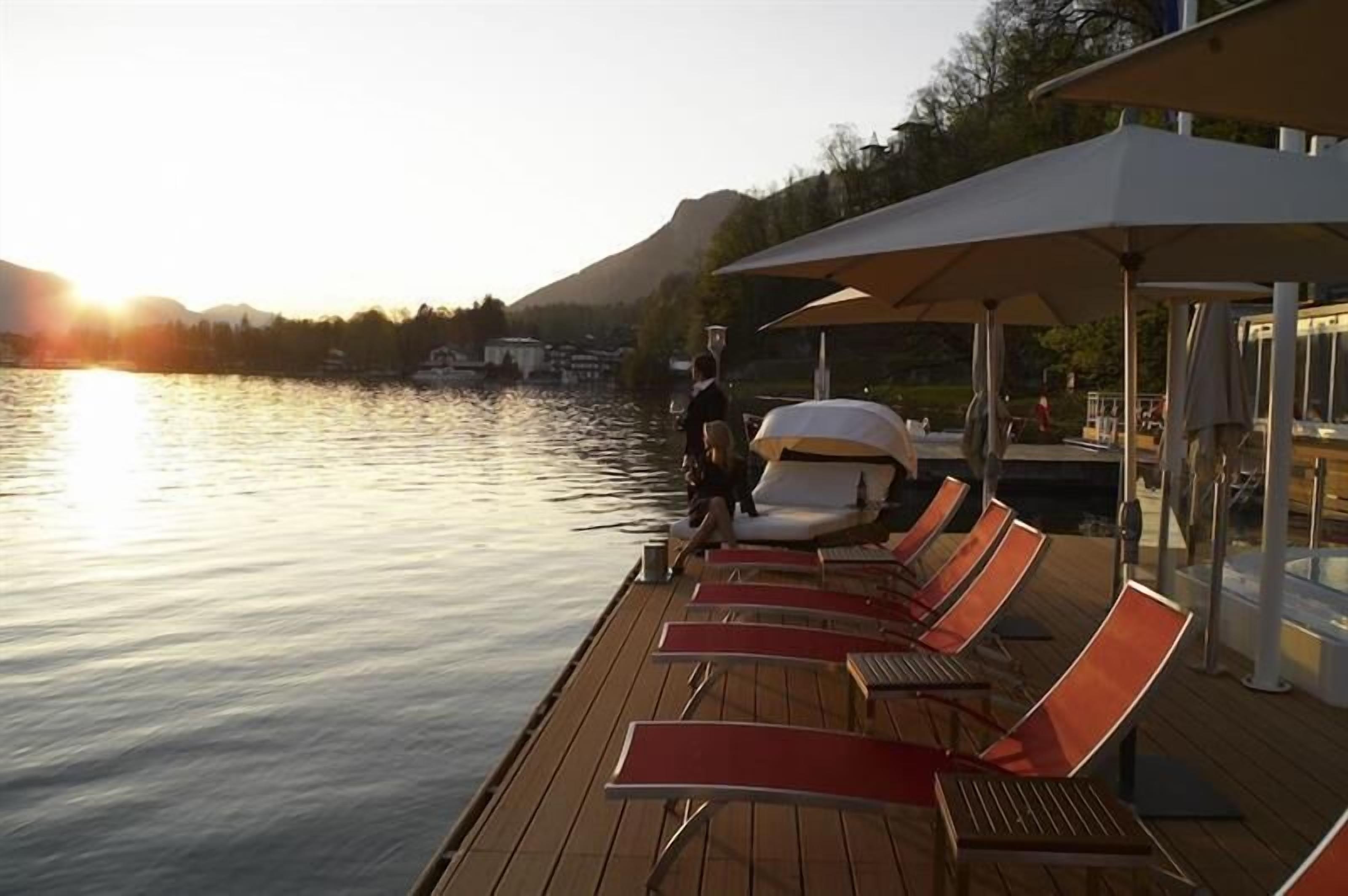 A wooden deck with sun loungers and umbrellas, overlooking a calm lake with a mountain in the background.