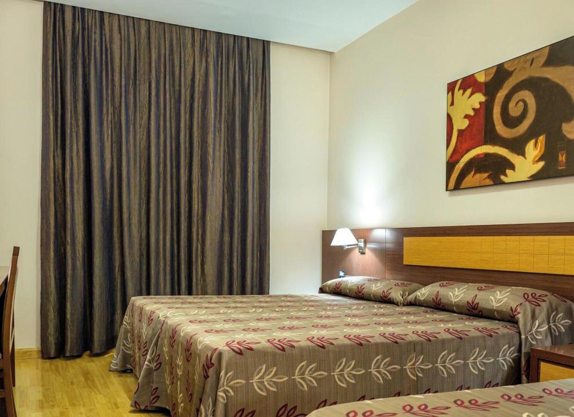 A hotel room with a bed, a nightstand, a lamp, and a large window with curtains.