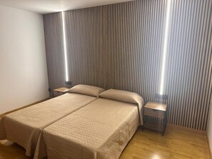 Un llit doble amb un edredó beix amb textura, dues taules de nit i una paret amb ratlles.