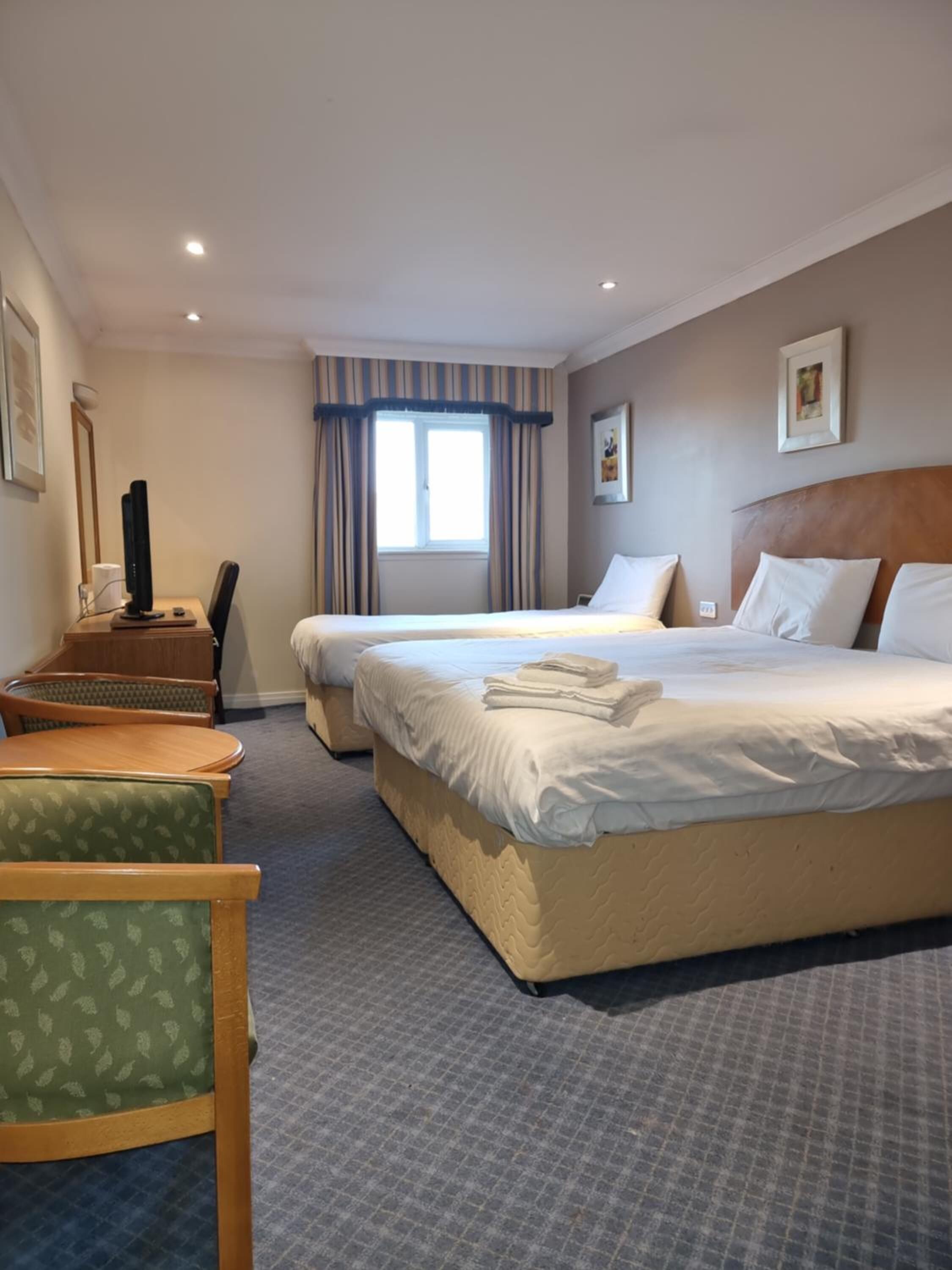 Habitación de hotel con una cama grande, un escritorio con computadora, una silla y una ventana con cortinas.
