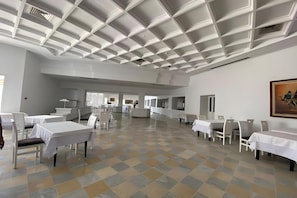 Un grand espace dédié à la salle à manger, avec des tables et des chaises blanches, un sol à carreaux et un plafond haut orné d’un motif en réseau.