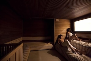 Una habitación tenuemente iluminada con paneles de madera y un amplio ventanal. Dos personas descansan en un banco.