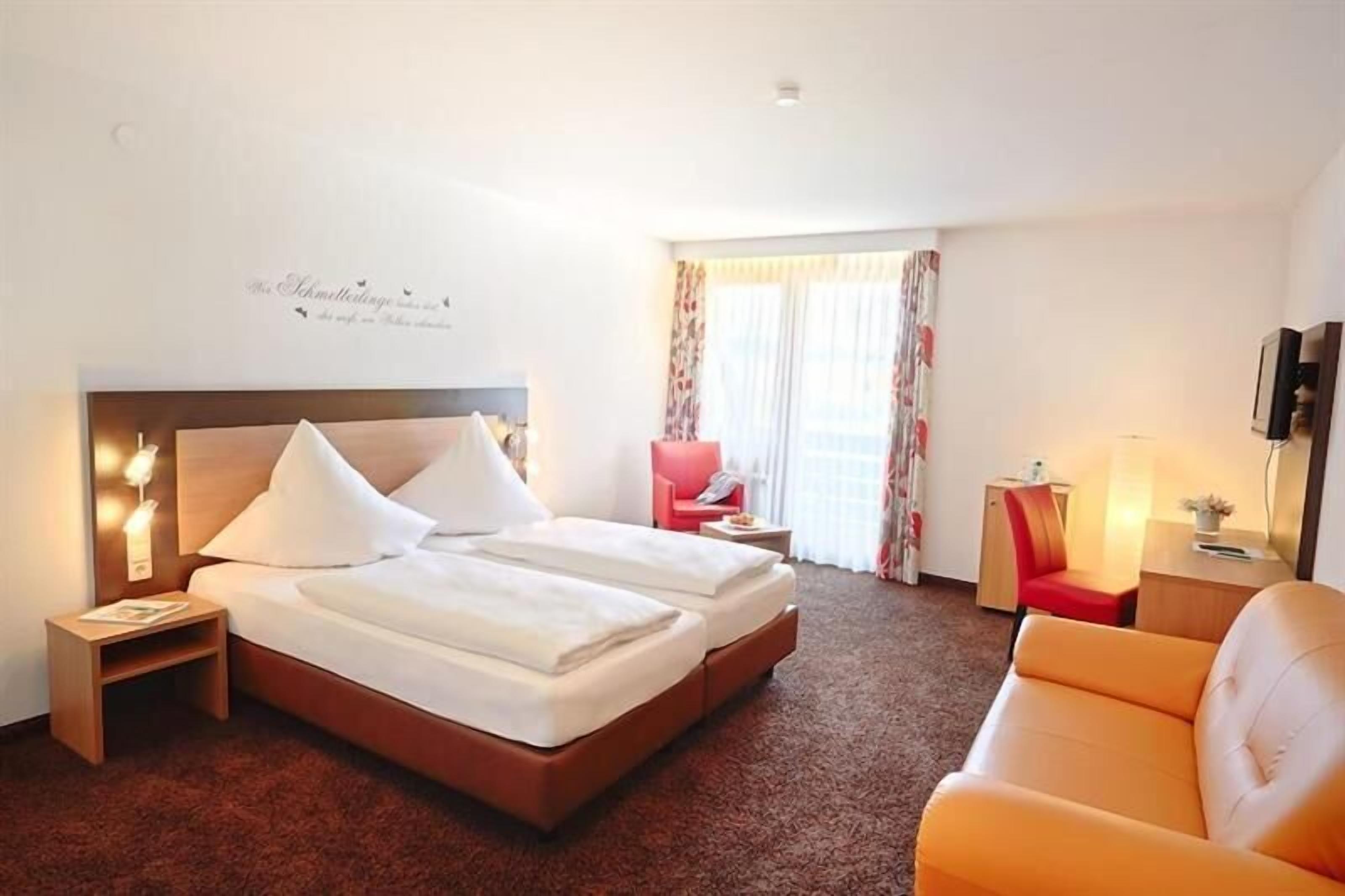 A hotel room with a double bed, a red chair, a sofa, a small table, and a TV.
