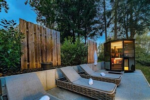 Een moderne buitenlounge met twee ligbedden en een kleine glazen constructie, omgeven door houten hekwerk en begroeiing.