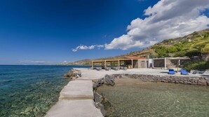 A coastal property with a clear view of the sea, a stone pathway leading to a pool, and a covered outdoor seating area with lounge chairs. - Lalibay Resort & Spa - Adults Only (Aegina)