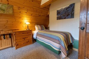 A cozy bedroom with a wooden headboard, a bedside lamp, a striped bedspread, and a mountain landscape painting on the wall. - Alaska Lakeside Cottage (Seward)