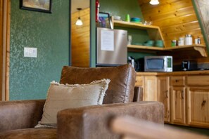 A cozy living area with a brown leather sofa and a cushion, a wooden kitchen with cabinets and appliances, and a green textured wall. - Alaska Lakeside Cottage (Seward)