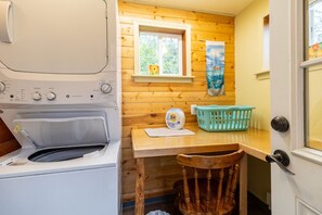 A compact laundry room with a washing machine, a small table with a basket, and a window with a view of greenery. - Alaska Lakeside Cottage (Seward)
