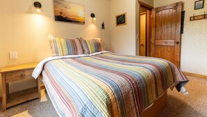 A bedroom with a bed, a nightstand, a wooden door, and a wall-mounted light fixture. - Alaska Lakeside Cottage (Seward)