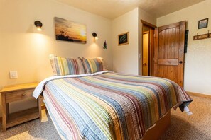 A bedroom with a bed, a nightstand, a wooden door, and a wall-mounted light fixture. - Alaska Lakeside Cottage (Seward)