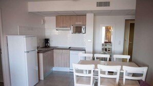 A compact kitchen with white appliances, a dining area with white chairs, and a visible microwave above the counter. - Résidence Nemea L'estacade (Capbreton)