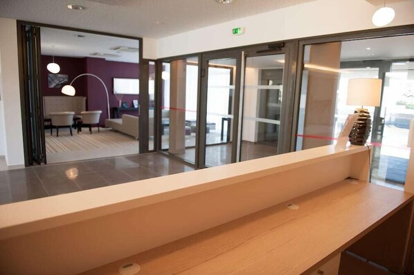 A reception area with a reception desk, glass doors, and a seating area with chairs and a lamp. - Résidence Nemea L'estacade (Capbreton)