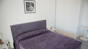 A neatly made bed with a purple blanket, a white bedside table with a lamp, and a small white radiator. - Résidence Nemea L'estacade (Capbreton)