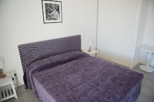 A neatly made bed with a purple blanket, a white bedside table with a lamp, and a small white radiator.