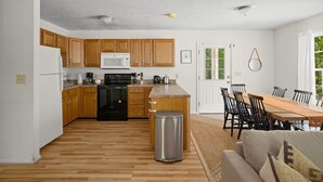 Un'ampia cucina con mobili in legno, un forno a microonde e un fornello. C'è un'area pranzo con tavolo e sedie, e un cestino nella parte centrale della stanza.