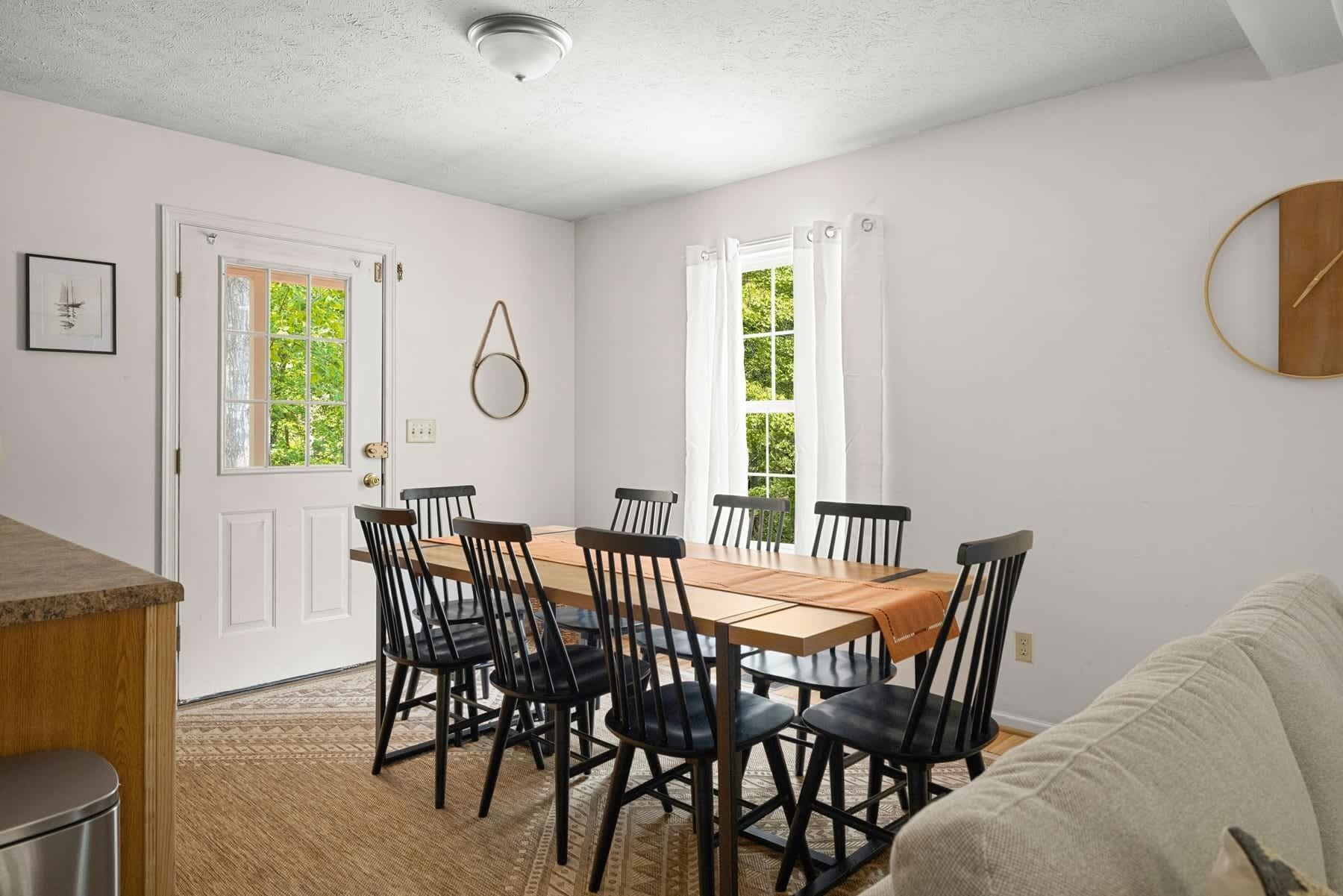 A dining area with a wooden table and black chairs, white walls, and windows with curtains.