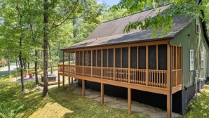 Eine Holzterrasse, an ein grünes Gebäude mit schrägem Dach angebaut, umgeben von Bäumen.