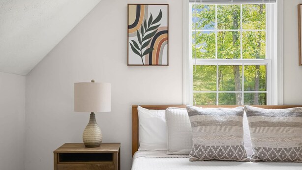 Una camera da letto con un comodino in legno, una lampada, un letto con biancheria da letto bianca e cuscini a motivi, una finestra vista alberi e un'opera d'arte a parete.