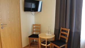 A small room with a wooden door, a TV mounted on the wall, two wooden chairs with black cushions, a round table with a glass pitcher and two glasses, and a dark curtain on the window. - North West Hotel & Restaurant (Hvammstangi)