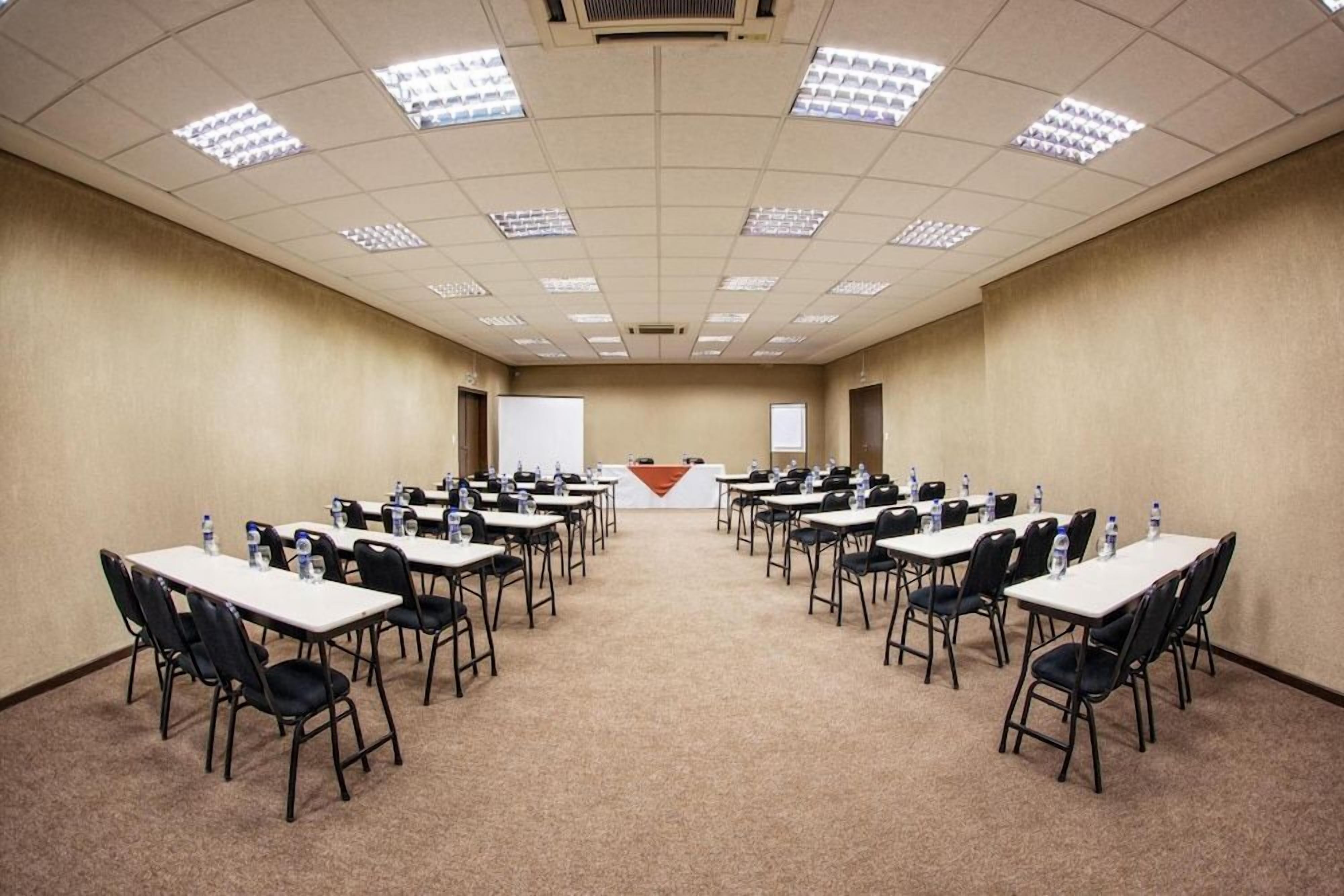 Uma sala de conferências com várias mesas e cadeiras dispostas em fileiras de frente para uma tela de projeção.