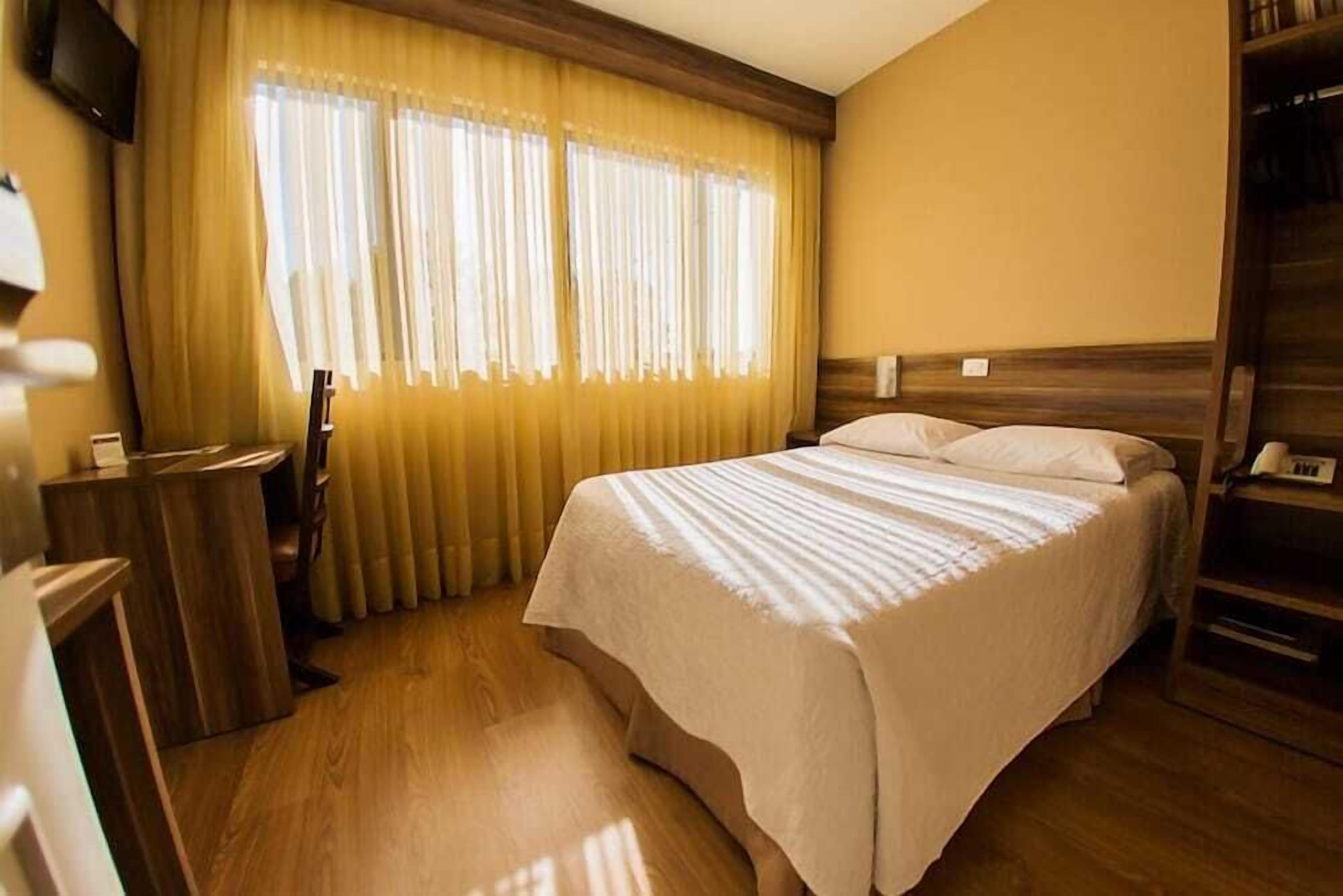 A hotel room with a bed, wooden headboard, and a window with curtains.