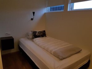 Ein kleines, ordentlich bezogenes Bett mit beiger Bettdecke und Kissen, ein Nachttisch und eine Wandleuchte.