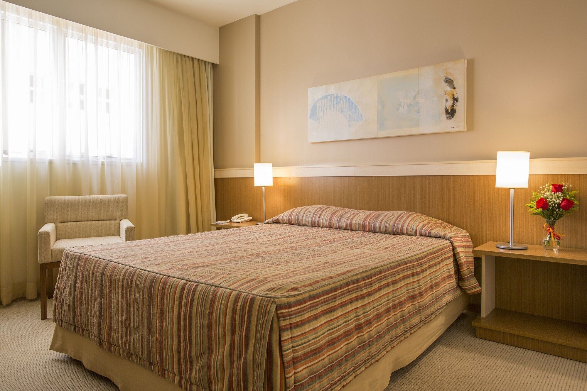 Ein Hotelzimmer mit einem großen Bett, einem Sessel, einem Nachttisch mit Lampe, einem Telefon und einem Bild an der Wand.