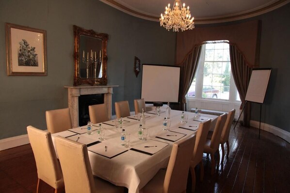 Una sala de conferencias con una larga mesa, sillas, una chimenea, un candelabro, un ventanal grande y una pizarra.