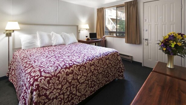 A hotel room with a large bed, two bedside lamps, a desk with a laptop, a window with curtains, and a vase with flowers on a dresser. - Westmark Inn Skagway (Skagway)